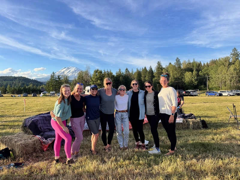 Eight GEA Alliance board members standing together in a green field, arms around each other, with a snow-capped mountain rising behind them under a blue sky