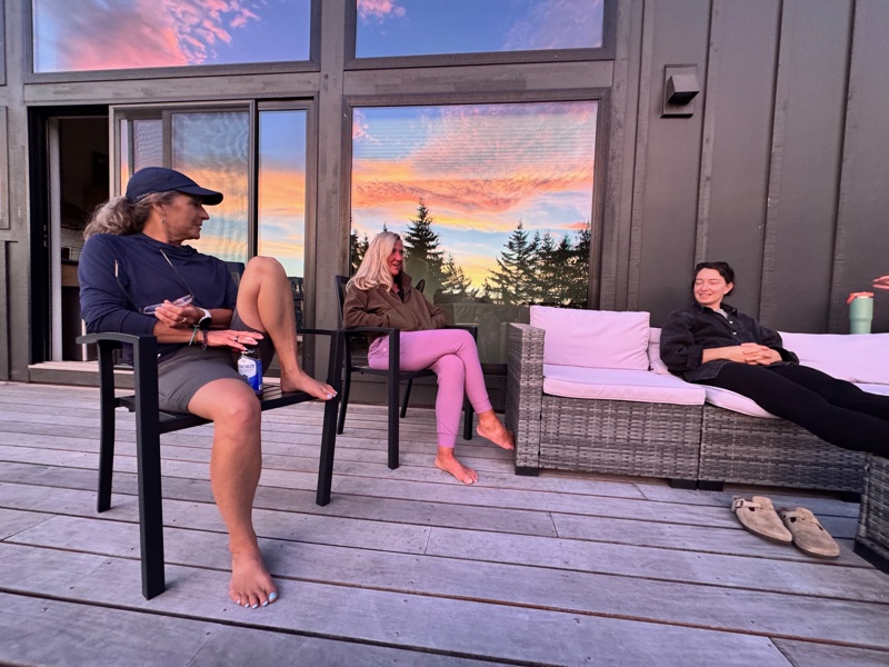 Three board members unwinding on the deck at golden hour — the Pacific Northwest sunset reflected brilliantly in the floor-to-ceiling windows of the retreat house