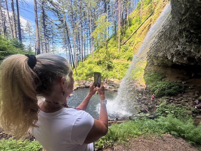 A board member photographs a tall waterfall with her phone, standing amid dense ferns and towering evergreens with the falls visible in the background