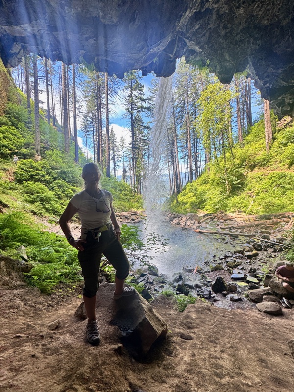 A board member stands silhouetted inside a mossy cave grotto, looking out at a tall waterfall cascading into a lush Pacific Northwest forest with brilliant green trees and a turquoise pool below