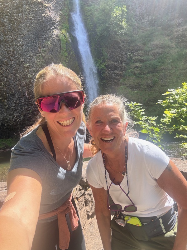 Two board members grinning at the camera in a selfie, with a waterfall tumbling down dark basalt cliffs and green ferns framing the scene behind them
