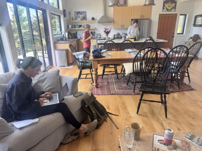Friday morning at the retreat house — three board members working in the open living-and-kitchen space, one on a laptop on the couch, two prepping coffee at the kitchen island