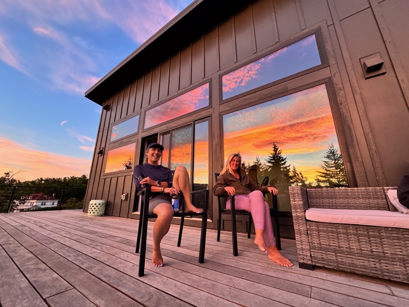 Two board members relaxing on the retreat house deck as a dramatic pink-and-orange sunset blazes across the sky and reflects in the tall windows behind them